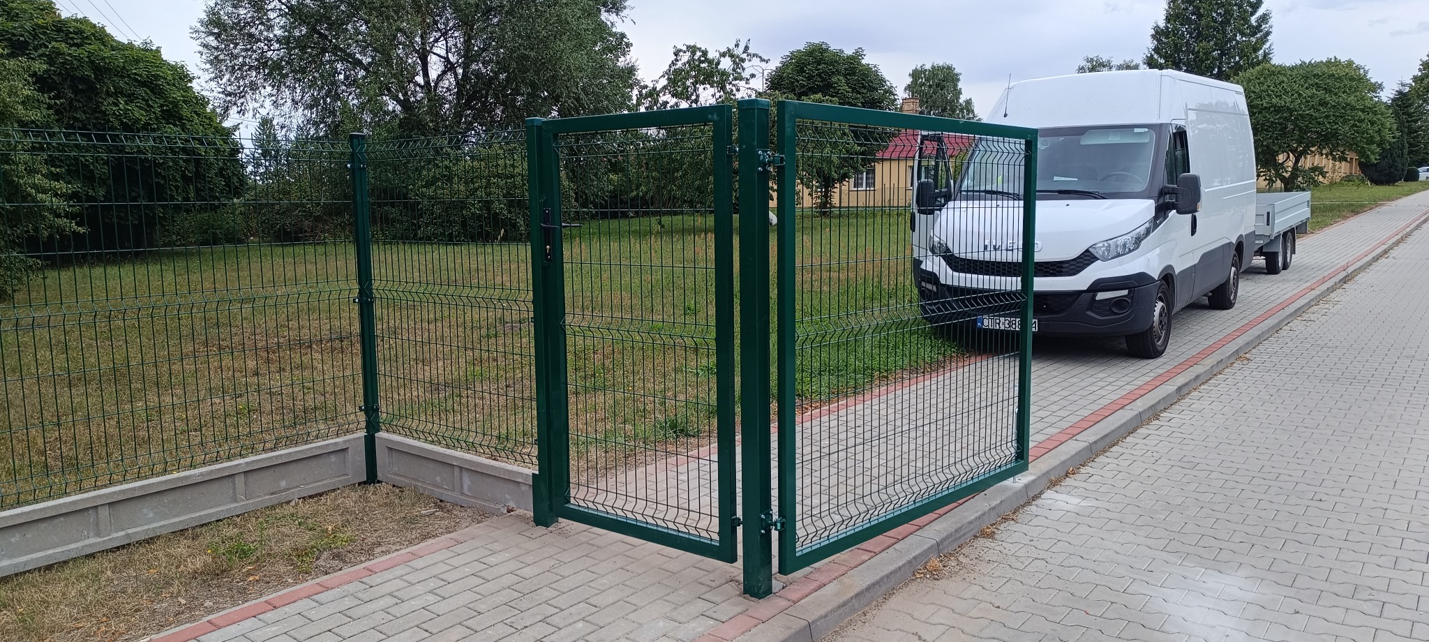 Zielona brama z siatki panelowej otwarta na posesję, z białym samochodem dostawczym w tle, widok z chodnika wyłożonego kostką.