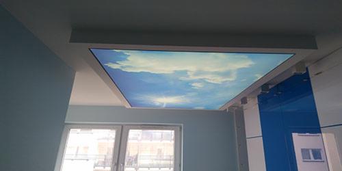 Nowoczesny sufit z podświetleniem LED imitującym niebo z chmurami, wkomponowany w białą ramę. Widok na okno i niebieskie elementy wystroju.