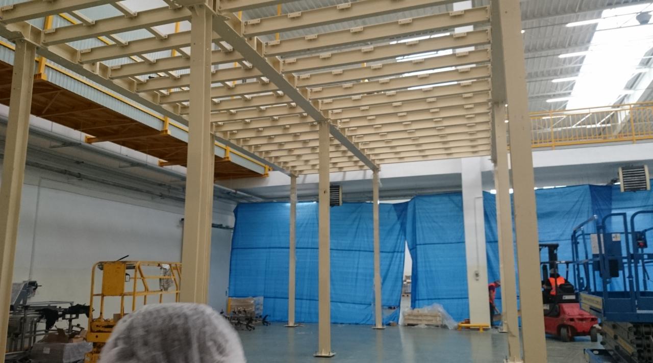 Wnętrze hali produkcyjnej z konstrukcją stalową pod sufitem, niebieskie płachty ochronne w tle i podnośniki koszowe, widoczny fragment żółtego wózka widłowego oraz osoba w stroju roboczym.