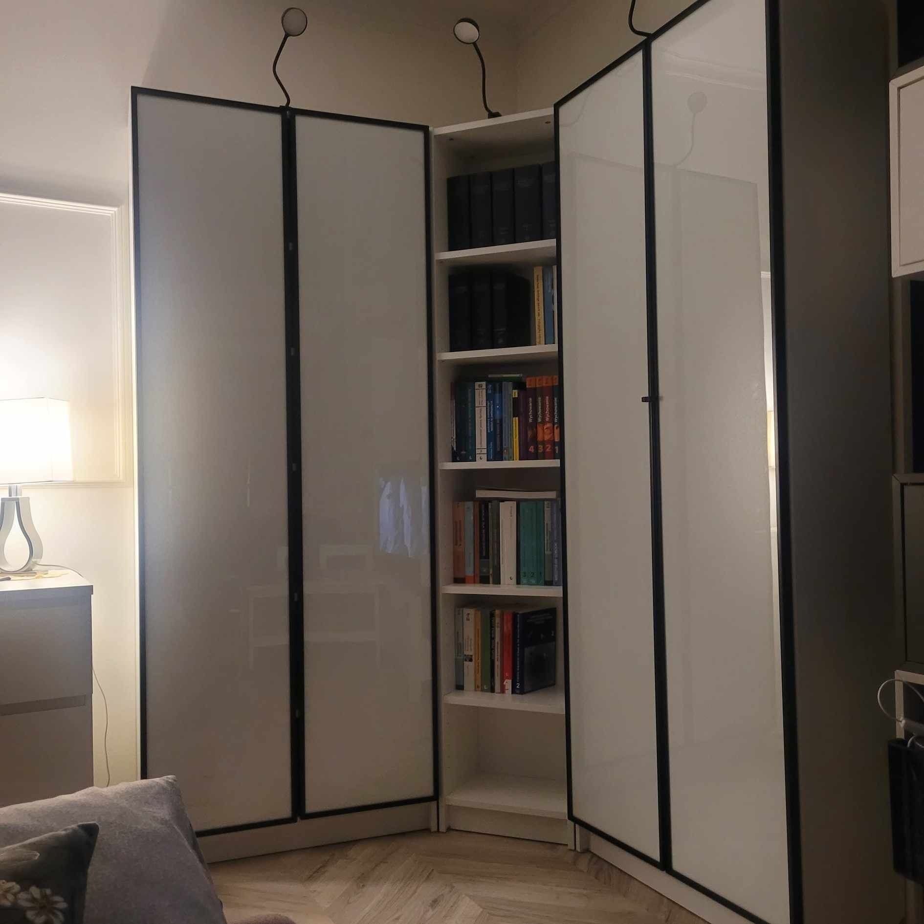 Kątowa szafa z białymi, przesuwanymi drzwiami z czarnymi ramami, oświetlona dwoma lampkami na giętkich ramionach, z otwartą półką wypełnioną książkami.