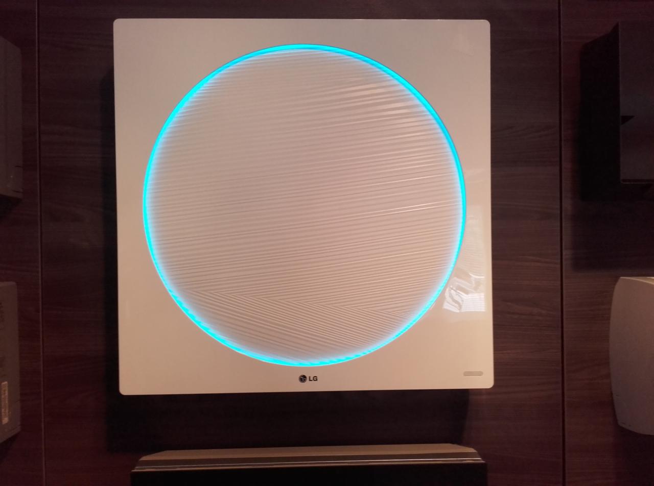 Nowoczesny klimatyzator LG w kształcie kwadratu, z okrągłym panelem i niebieskim podświetleniem, zamontowany na ścianie z drewnianą okładziną.