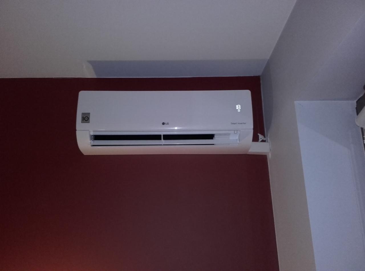 Biała klimatyzacja LG Smart Inverter zamontowana na bordowej ścianie, z widocznym wyświetlaczem temperatury i fragmentem białego sufitu.