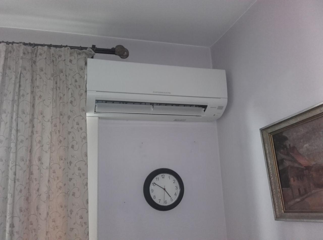 Biała klimatyzacja Mitsubishi Electric na ścianie, pod nią okrągły zegar i obraz w ramie. Zasłona z ornamentem widoczna po lewej stronie.