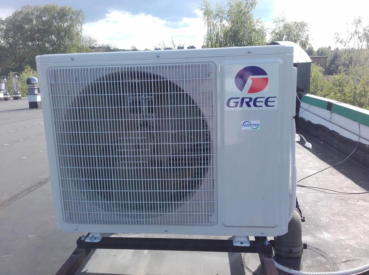 Zewnętrzna jednostka klimatyzacji Gree Inverter, zamontowana na płaskim dachu z widocznymi elementami instalacji. Ujęcie z boku, z zielenią w tle.