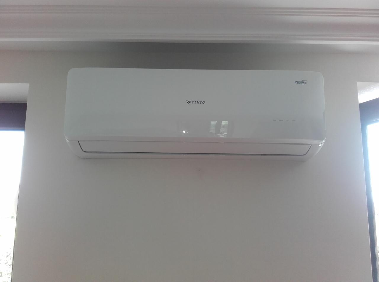 Zamontowana, biała klimatyzacja Rotenso na jasnej ścianie, widoczna z przodu. Obok okno z widocznym światłem słonecznym. Minimalistyczny design i czysty montaż.