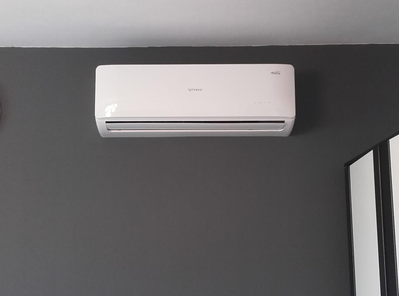 Biała klimatyzacja Rotenso na szarej ścianie w Wieliczce. Widoczne delikatne odbicia światła i fragment szafy w tle. Nowoczesny design, montaż w domu.