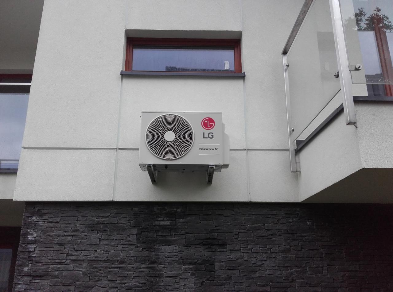 Zamontowany na elewacji budynku klimatyzator LG Inverter V, widoczny z boku, na tle okna i szklanej balustrady balkonu.