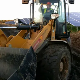 Żółta ładowarka Zeppelin z operatorem w kasku i kamizelce, praca w terenie z panelami słonecznymi w tle. Błoto na oponach i łyżce.