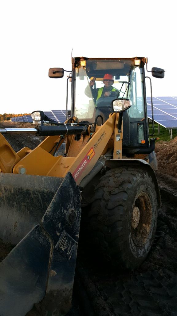 Żółta ładowarka Zeppelin z operatorem w kasku i kamizelce, praca w terenie z panelami słonecznymi w tle. Błoto na oponach i łyżce.