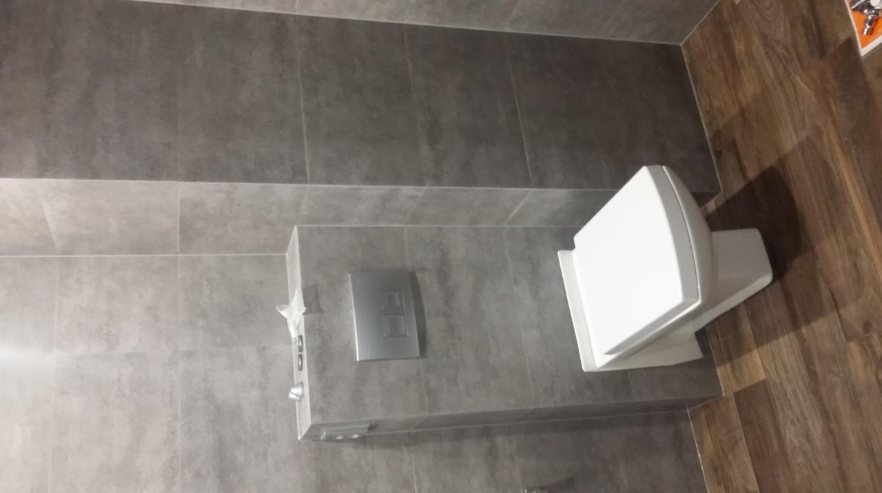 Nowoczesna toaleta podwieszana z szarą, matową okładziną ścienną i drewnopodobną podłogą. Widoczny chromowany przycisk spłuczki.