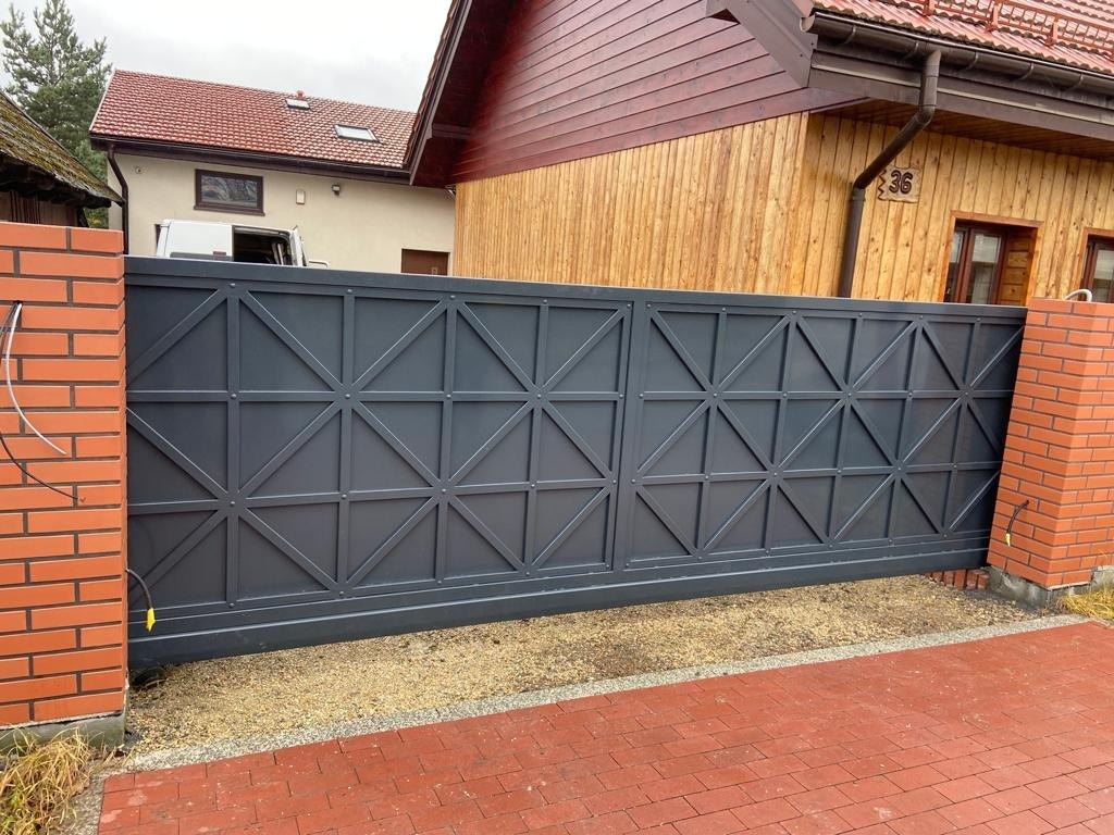 Szara, przesuwna brama wjazdowa z geometrycznym wzorem kraty, zamontowana między ceglanymi słupkami, na tle domu z drewnianą elewacją.