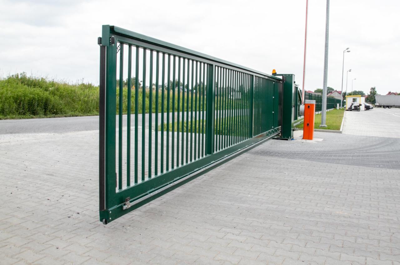 Otwarta, przesuwna brama przemysłowa w kolorze ciemnej zieleni, z widocznym mechanizmem zębatkowym, na tle betonowej nawierzchni i zieleni.