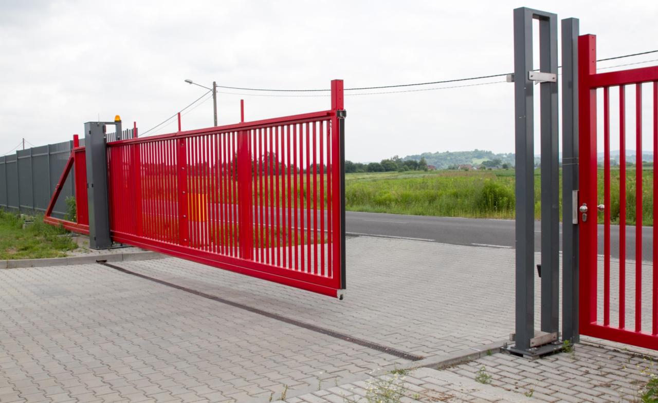 Czerwona przesuwna brama wjazdowa z pionowymi szczeblami, częściowo otwarta, z szarą konstrukcją nośną i widocznym mechanizmem automatycznym, na tle kostki brukowej i krajobrazu.