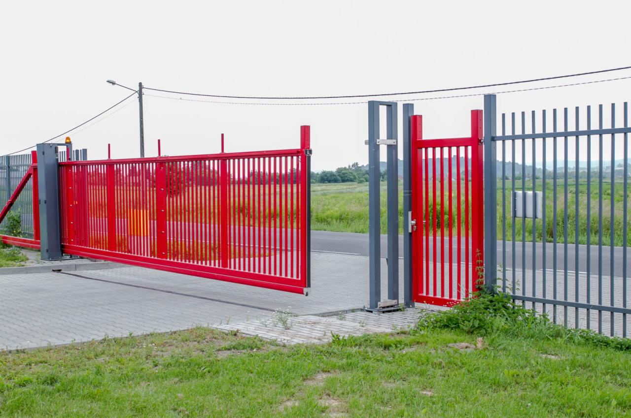 Częściowo otwarta, przesuwna, czerwona brama wjazdowa z furtką w szarym ogrodzeniu panelowym, widok z zewnątrz posesji.