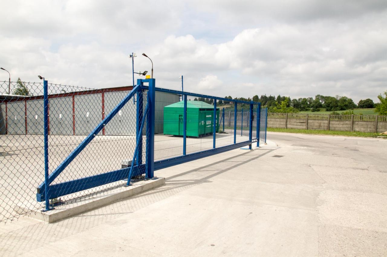 Niebieska brama przesuwna z siatki na betonowej posadzce, w tle szary blaszak i zielony kontener na śmieci, widok z boku.