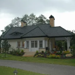 Elegancki dom jednorodzinny z ciemną dachówką, jasną elewacją i białymi okiennicami, otoczony zadbanym ogrodem z kolorowymi kwiatami i brukowaną ścieżką.