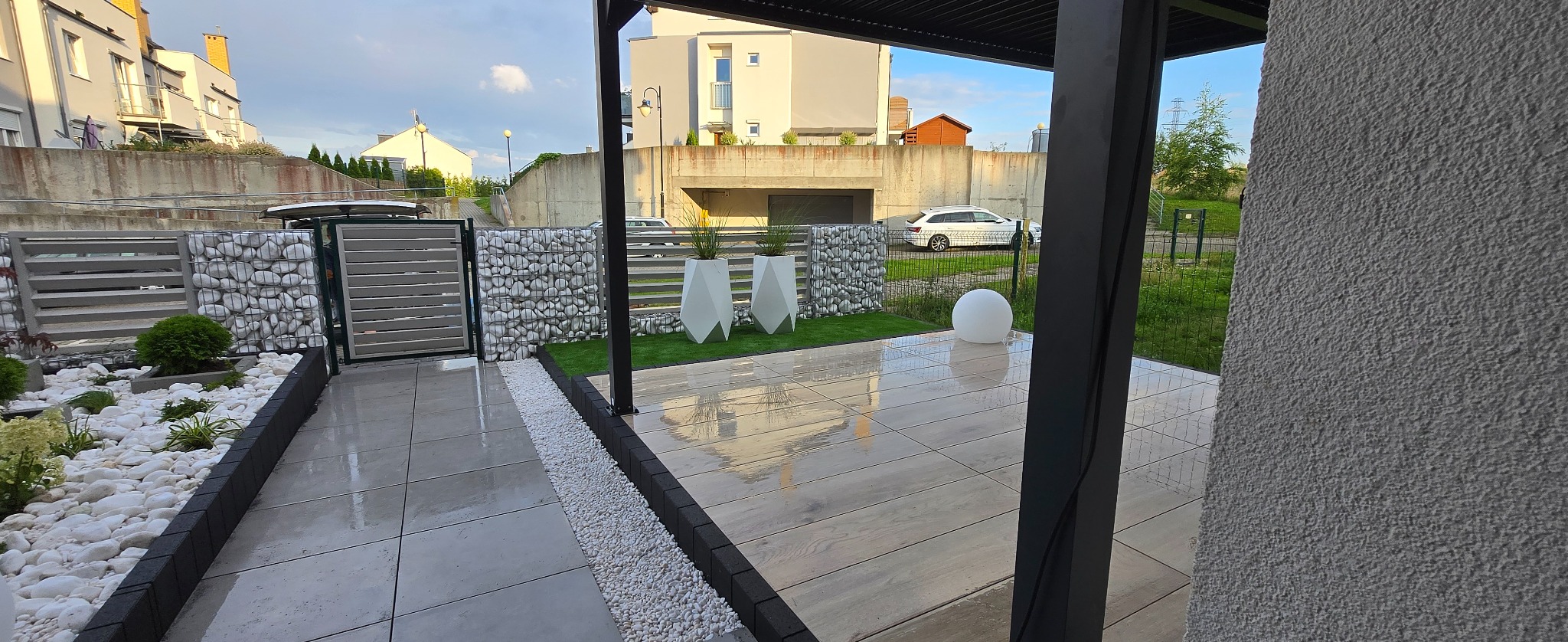 Nowoczesny taras z jasnymi płytami, białymi kamieniami i gabionami. Minimalistyczne donice i oświetlenie kuliste. Widok na dom i zieleń. Po deszczu, mokra nawierzchnia.