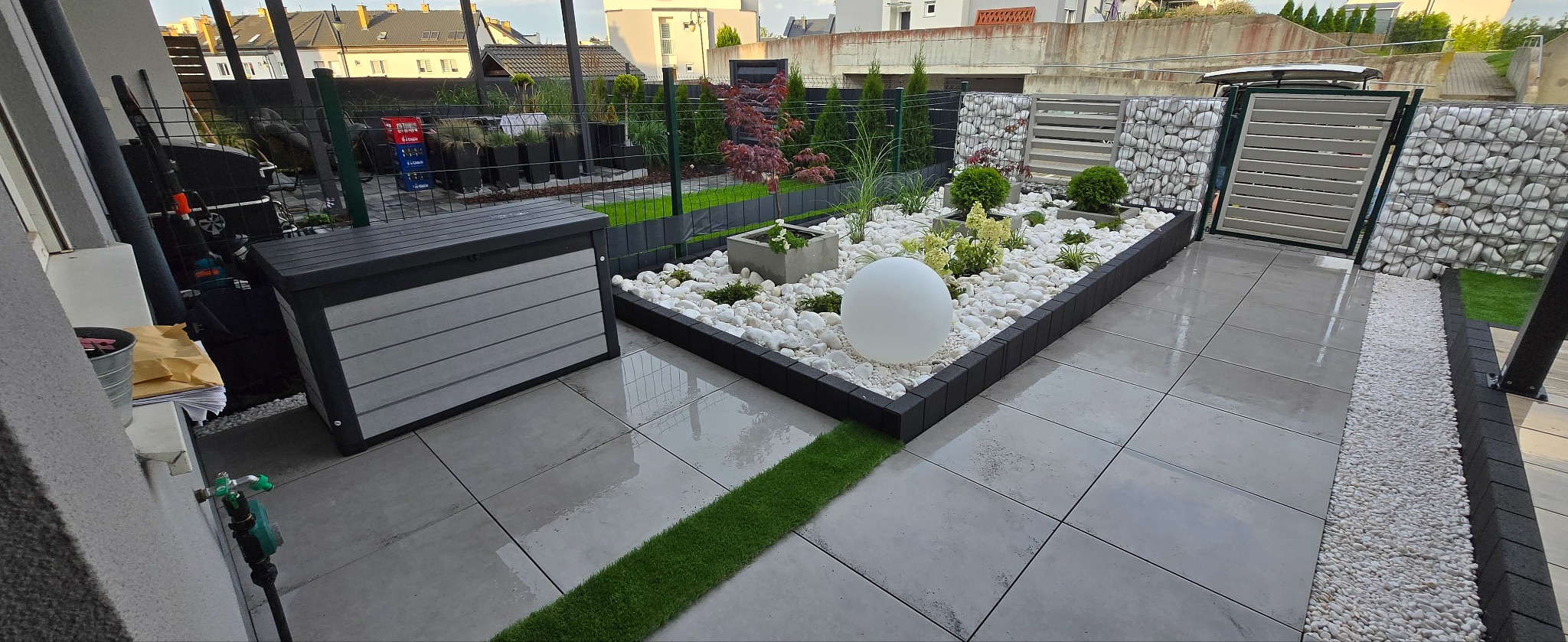 Nowoczesny ogród z białymi kamieniami, roślinami w donicach i oświetleniem sferycznym. Szare płytki tarasowe i zielony pas trawy tworzą elegancką przestrzeń.