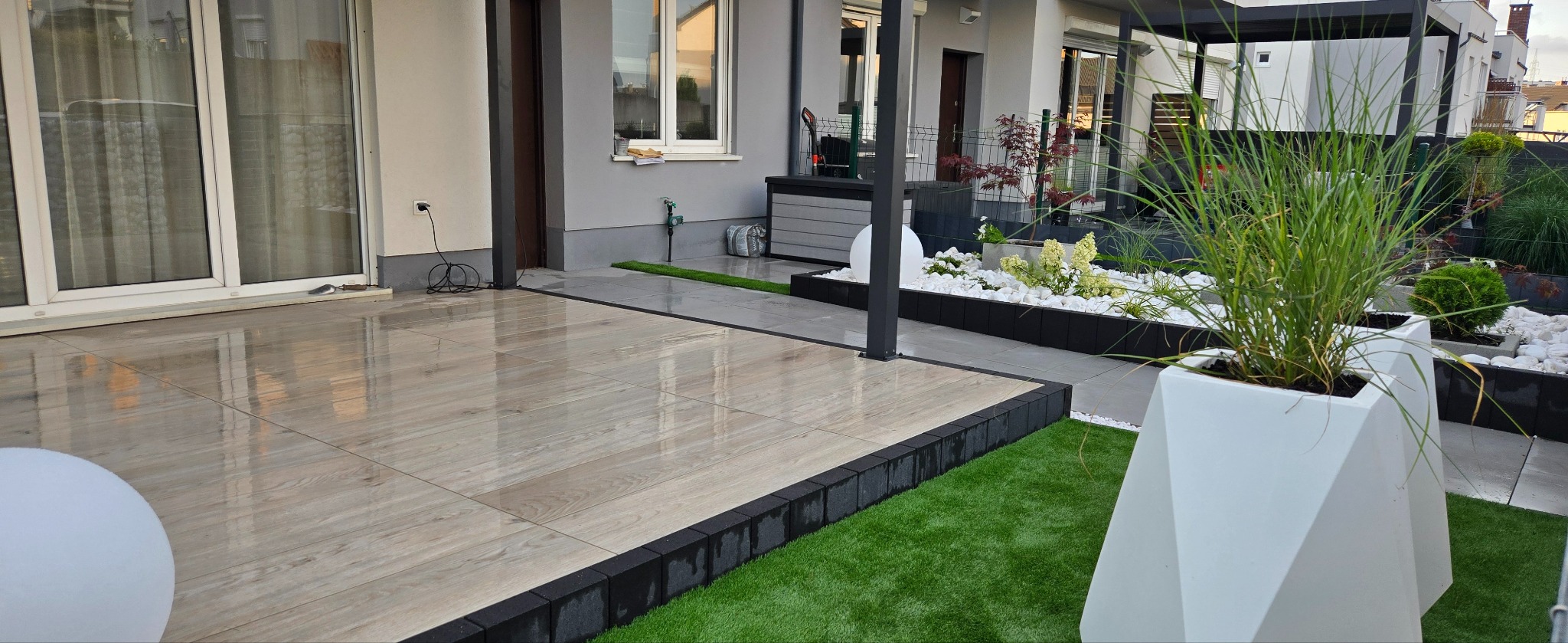 Nowoczesny taras z drewnopodobną płytką, obrzeżem z kostki i sztuczną trawą. W tle pergola i roślinność ozdobna w białych donicach.