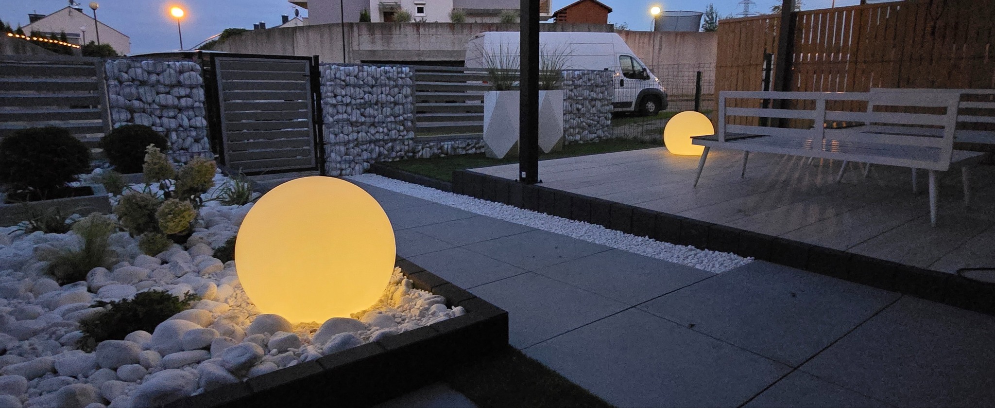 Oświetlony ogród z kulistymi lampami, białymi kamieniami i gabionami w tle. Nowoczesny design ogrodu z elementami dekoracyjnymi i meblami ogrodowymi.