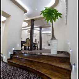 Stylowe schody z drewna prowadzą do eleganckiej jadalni. Dekoracyjny dywan, oświetlenie punktowe i roślina doniczkowa dodają wnętrzu charakteru.