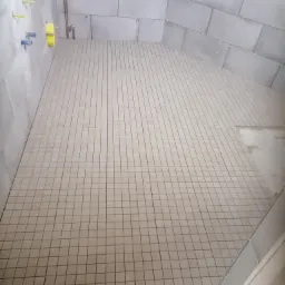 Świeżo ułożona, jasna mozaika podłogowa w szarej łazience, widoczne instalacje wodne na ścianie, płytki ścienne z krzyżykami dystansowymi.