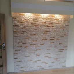 Ściana wykończona dekoracyjną okładziną z imitacji kamienia, podświetlona od góry, z widocznymi panelami podłogowymi i fragmentem drzwi.