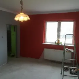 Pomieszczenie po malowaniu ścian na czerwono i szaro, widoczna drabina, grzejnik zabezpieczony folią i lampa sufitowa.