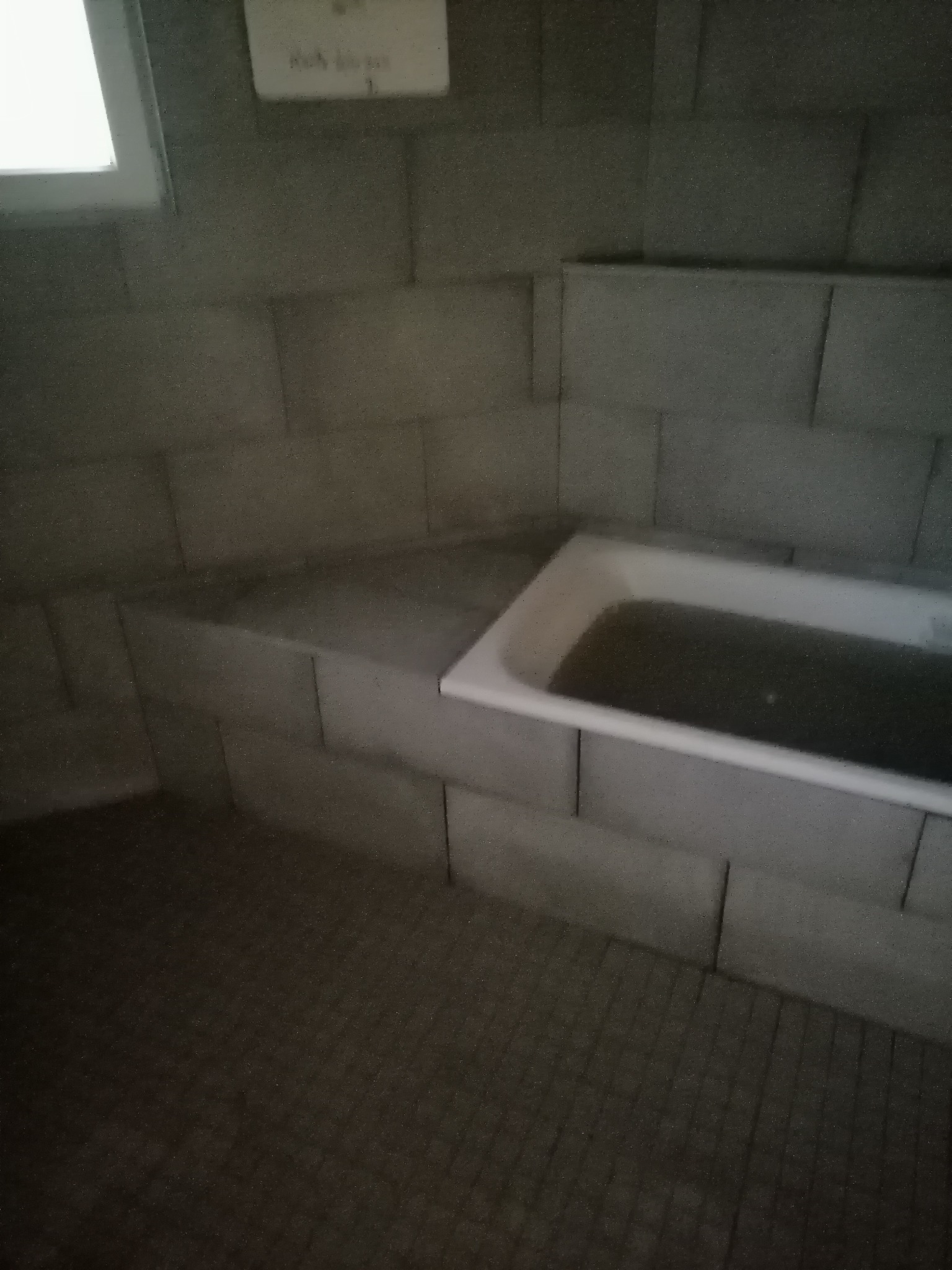 Szara łazienka z wanną wypełnioną ciemną wodą, widok z góry, ściany wyłożone płytami imitującymi cegły, małe okno w górnym lewym rogu, słabe oświetlenie.