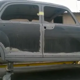 Klasyczny samochód w trakcie renowacji, z widocznymi śladami szpachli i przygotowania do lakierowania, umieszczony na mobilnej platformie warsztatowej.