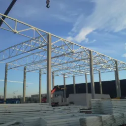 Wznoszenie stalowej hali: szkielet konstrukcji z dźwigiem w tle, płyty warstwowe składowane na placu budowy, błękitne niebo z chmurami nad budową.