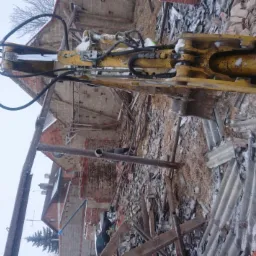 Żółty hydrauliczny młot wyburzeniowy na tle gruzowiska z cegieł i resztek konstrukcji budynku w zimowej scenerii, z widocznym fragmentem niebieskiej plandeki.