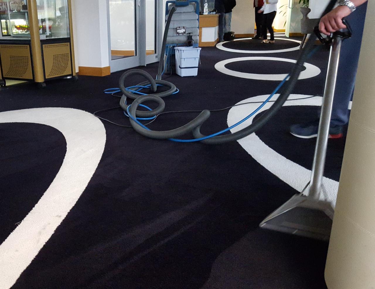 Czyszczenie wykładziny w biurze za pomocą profesjonalnego odkurzacza piorącego z metalową końcówką na tle wzorzystej, ciemnej podłogi.