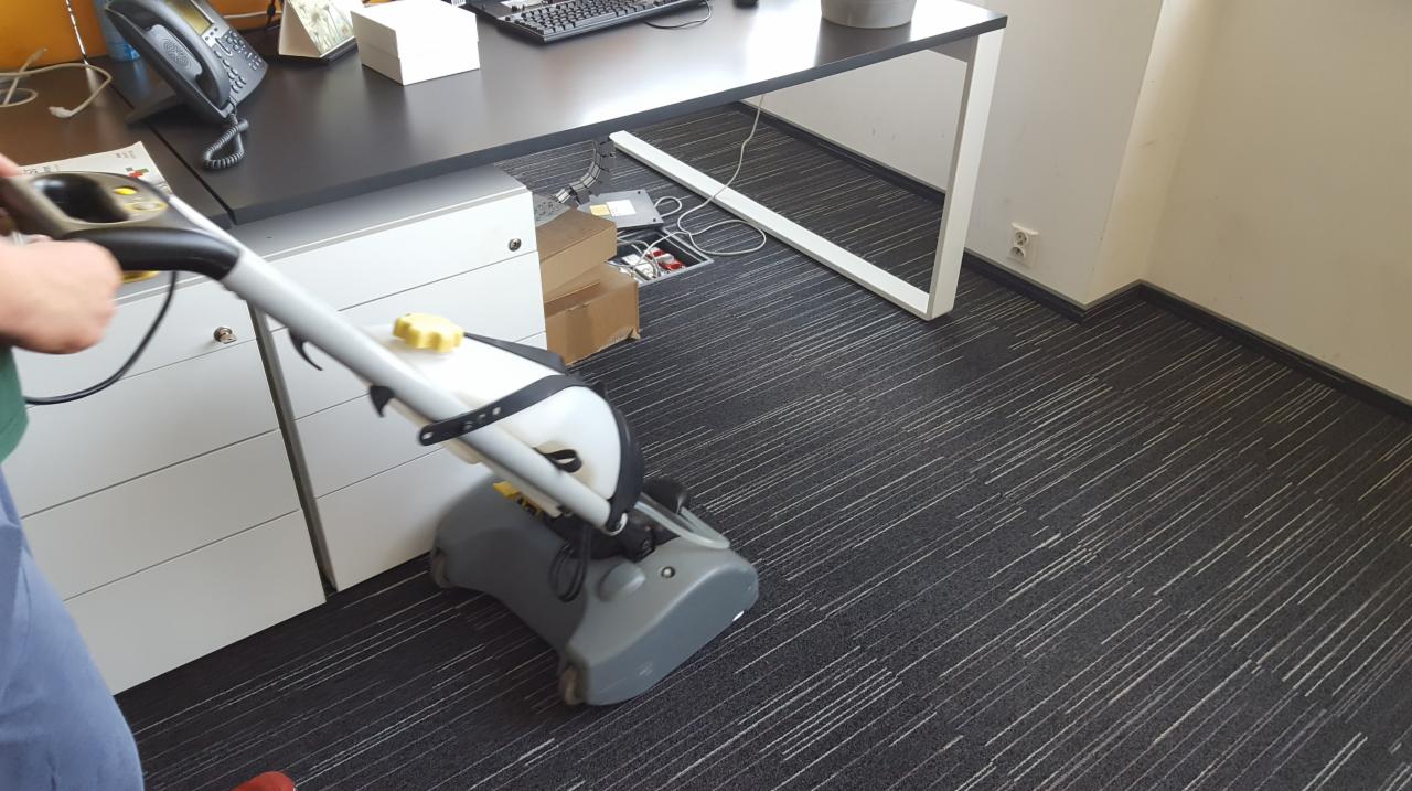 Czyszczenie wykładziny w biurze za pomocą profesjonalnego odkurzacza piorącego. Widoczne biurko z szafkami i otwarty kanał kablowy w podłodze.