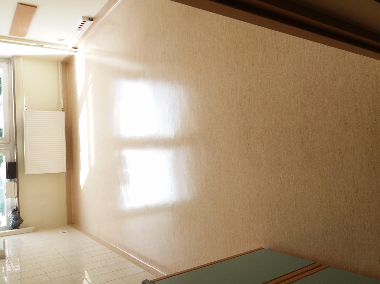 Wnętrze biura po odnowieniu: świeżo pomalowana ściana w odcieniach beżu z widocznymi refleksami światła słonecznego, jasna podłoga z płytek, grzejnik pod oknem i fragment mebla w rogu.