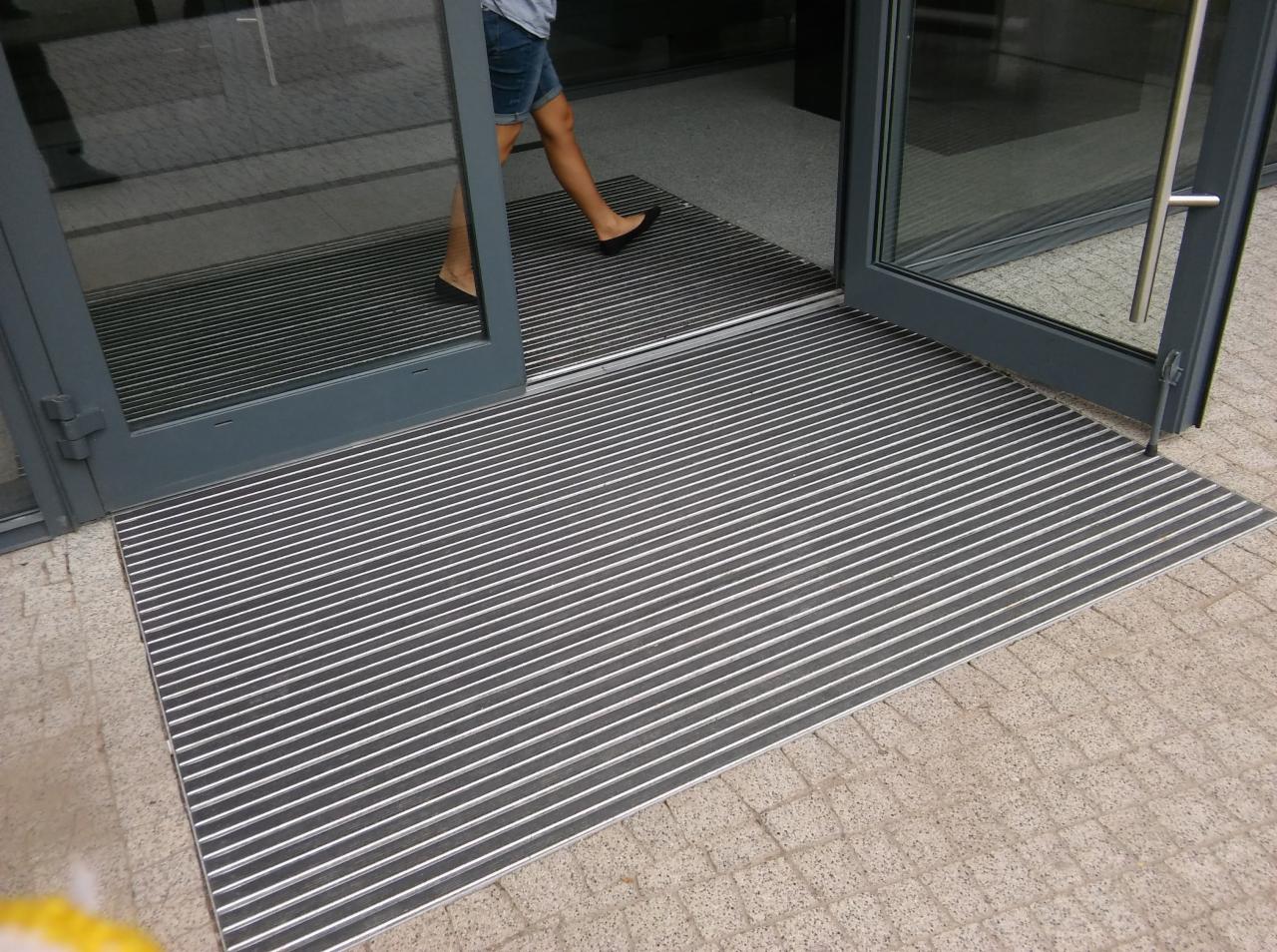 Otwarty szary szklany drzwi z metalową wycieraczką w paski, osoba w dżinsowych szortach wchodzi do budynku.