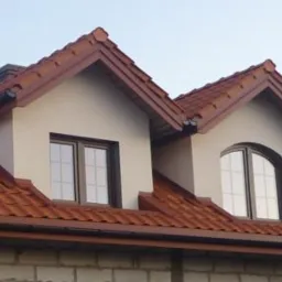 Dom w stanie surowym z nowym, brązowym dachem z dachówki ceramicznej i rynnami. Widoczne okna dachowe.