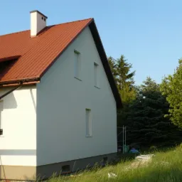 Świeżo otynkowana elewacja domu jednorodzinnego z czerwoną dachówką, widoczne rusztowanie i trawa w tle.