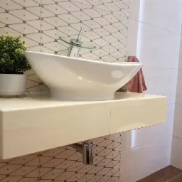 Biała, owalna umywalka nablatowa z chromowaną baterią, ustawiona na podwieszanej szafce z beżowych płytek, na ścianie płytki w geometryczny wzór, obok doniczka z zieloną rośliną.