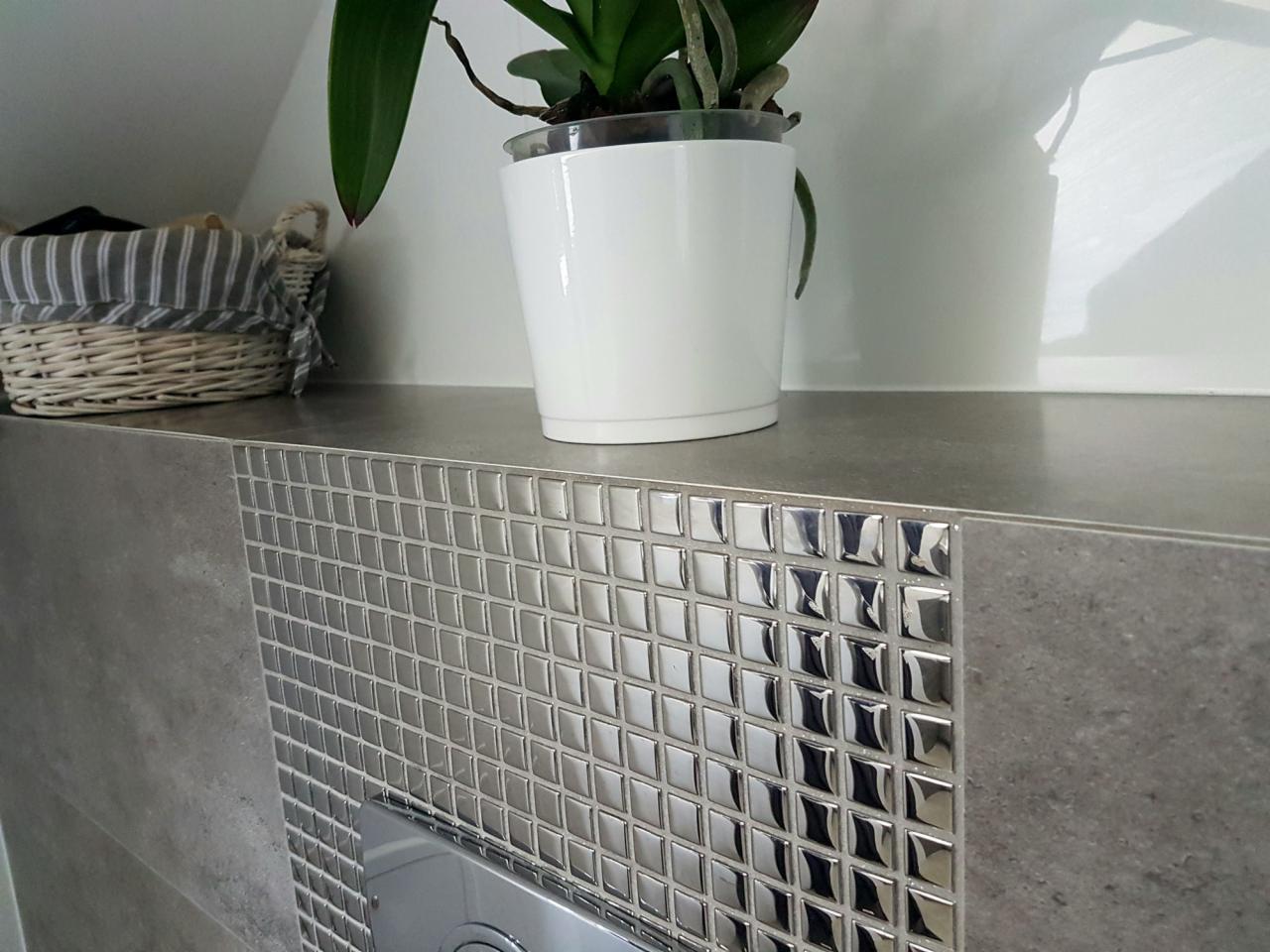 Błyszczące, srebrne mozaiki kwadratowe zdobią ścianę w łazience nad ukrytym zbiornikiem toalety, na półce stoi biała doniczka z zieloną rośliną, a obok wiklinowy koszyk z tkaniną w paski.