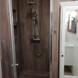 Nowoczesna kabina prysznicowa z ciemnobrązowymi płytkami imitującymi drewno, chromowana armatura i odpływ liniowy w podłodze, obok biała szafka z lustrem odbijającym umywalkę