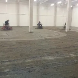 Wyrównywanie wylewki w hali przemysłowej za pomocą mechanicznych zacieraczek. Widoczne ślady po maszynach i kolumny konstrukcyjne.