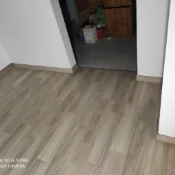 Ukończona podłoga z płytek imitujących drewno ułożonych w jodełkę w pokoju na poddaszu, widoczne białe ściany i otwarte drzwi do zagraconego pomieszczenia.