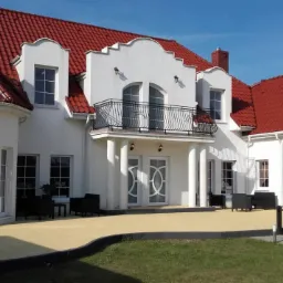 Elewacja dużego, białego domu z czerwonym dachem, widoczne okna z podziałami i dwuskrzydłowe drzwi wejściowe z dekoracyjnymi przeszkleniami.