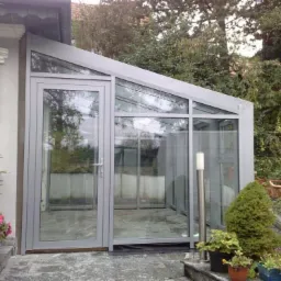 Szklana oranżeria z aluminiową ramą, przylegająca do domu, z widokiem na ogród, oświetlona lampą zewnętrzną.