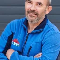 Uśmiechnięty mężczyzna z brodą w niebieskiej bluzie z logo firmy budowlanej na tle szarej ściany.