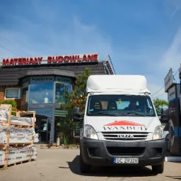 Biały samochód dostawczy firmy Waxbud Iveco na tle budynku sklepu z materiałami budowlanymi i pokryciami dachowymi, palety ze styropianem i dachówkami przed budynkiem.