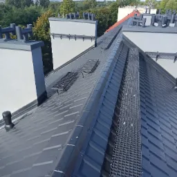 Nowy, grafitowy dach z blachy na budynku wielorodzinnym, widoczne kominy z czarnymi zakończeniami i metalowe pomosty komunikacyjne na dachu.