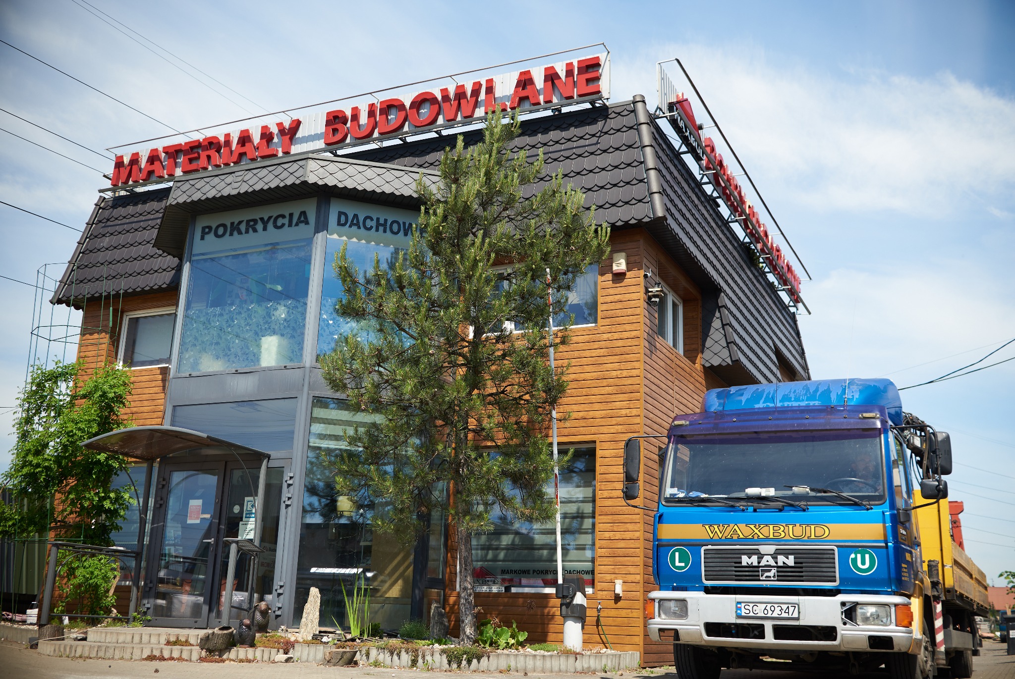 Budynek sklepu z materiałami budowlanymi z napisem 'Materiały Budowlane' i 'Pokrycia Dachowe' oraz zaparkowana ciężarówka MAN z napisem 'WAXBUD'.