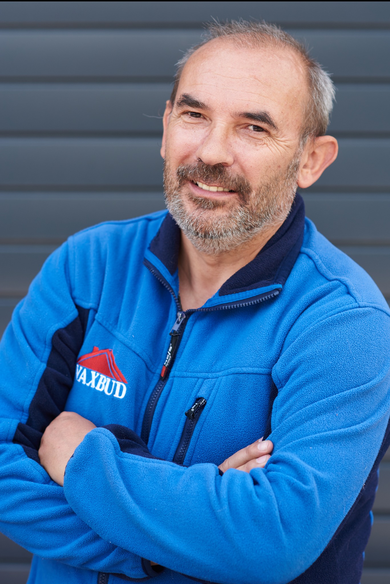 Uśmiechnięty mężczyzna z brodą w niebieskiej bluzie z logo firmy budowlanej na tle szarej ściany.